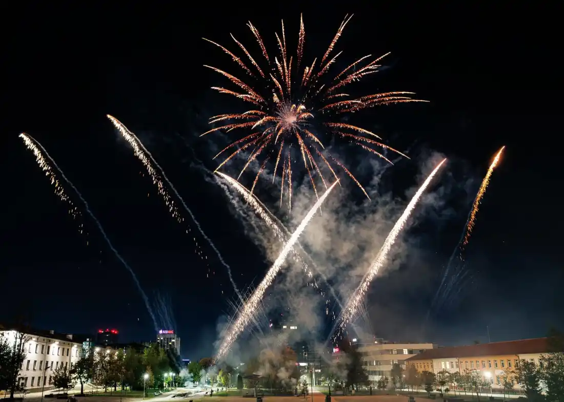 Pyrotechnics over a city square at night, the surrounding buildings lit up